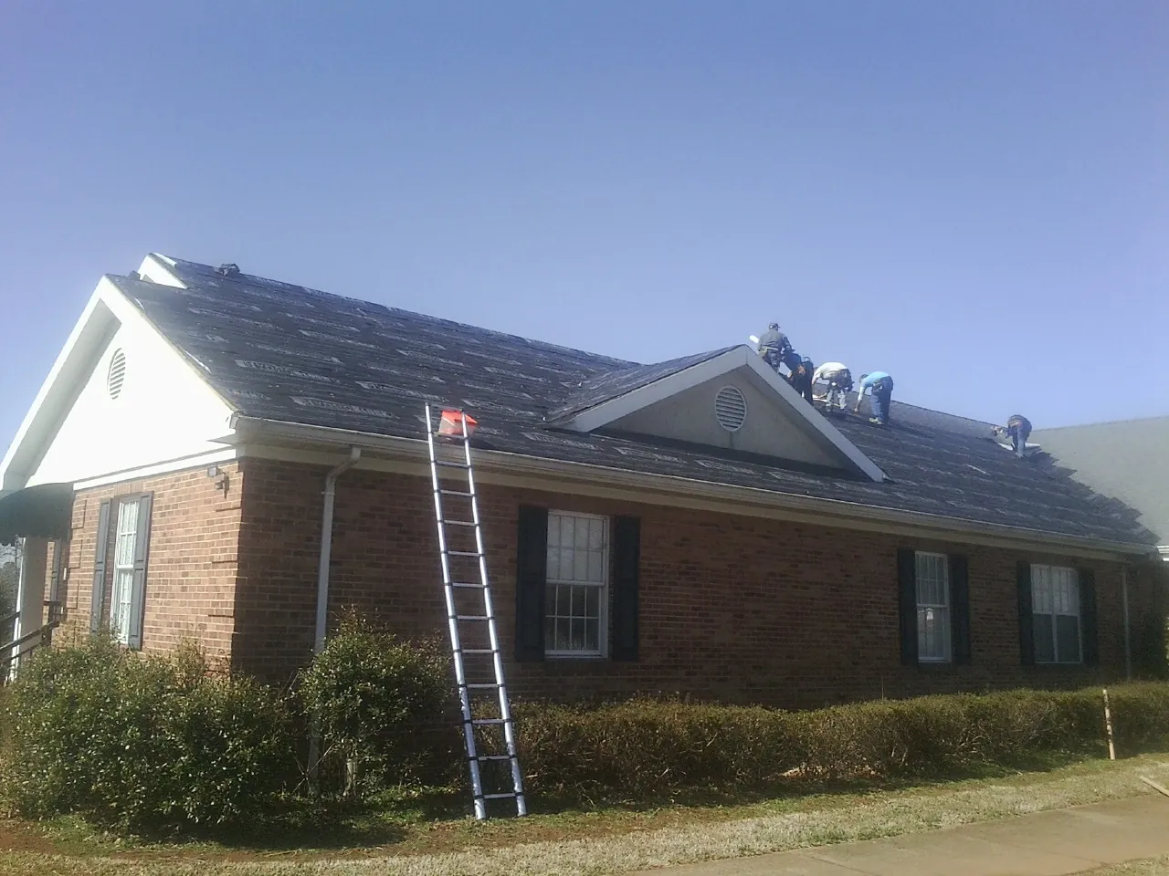 Crew tearing shingles off a brick ranch roof with a ladder set — Emergency Roof Repair work by our crew in Port Gibson