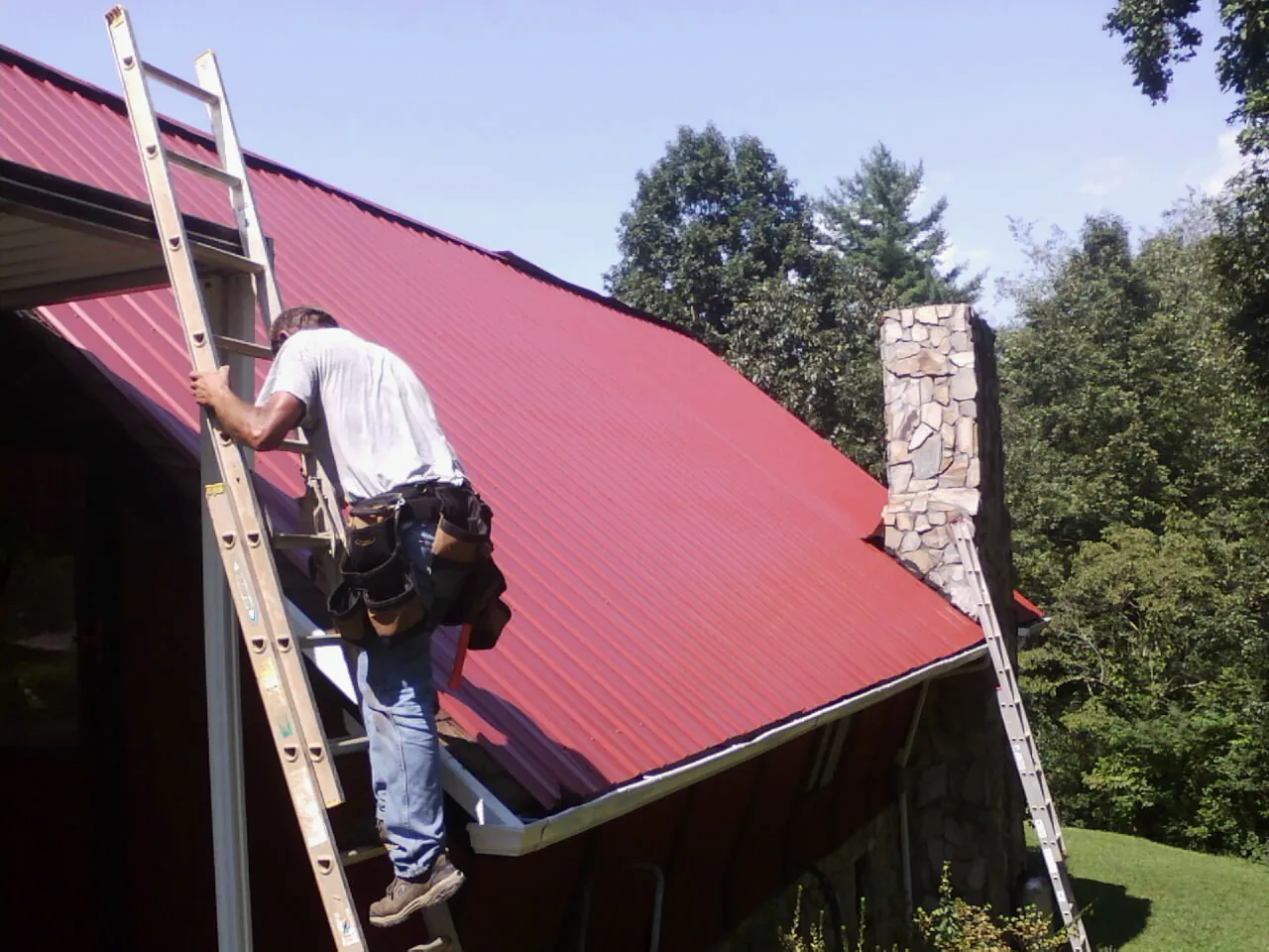 Roofer on a ladder beside a red metal roof and stone chimney — Soffit & Fascia Repair by Roofing Brothers in Port Gibson